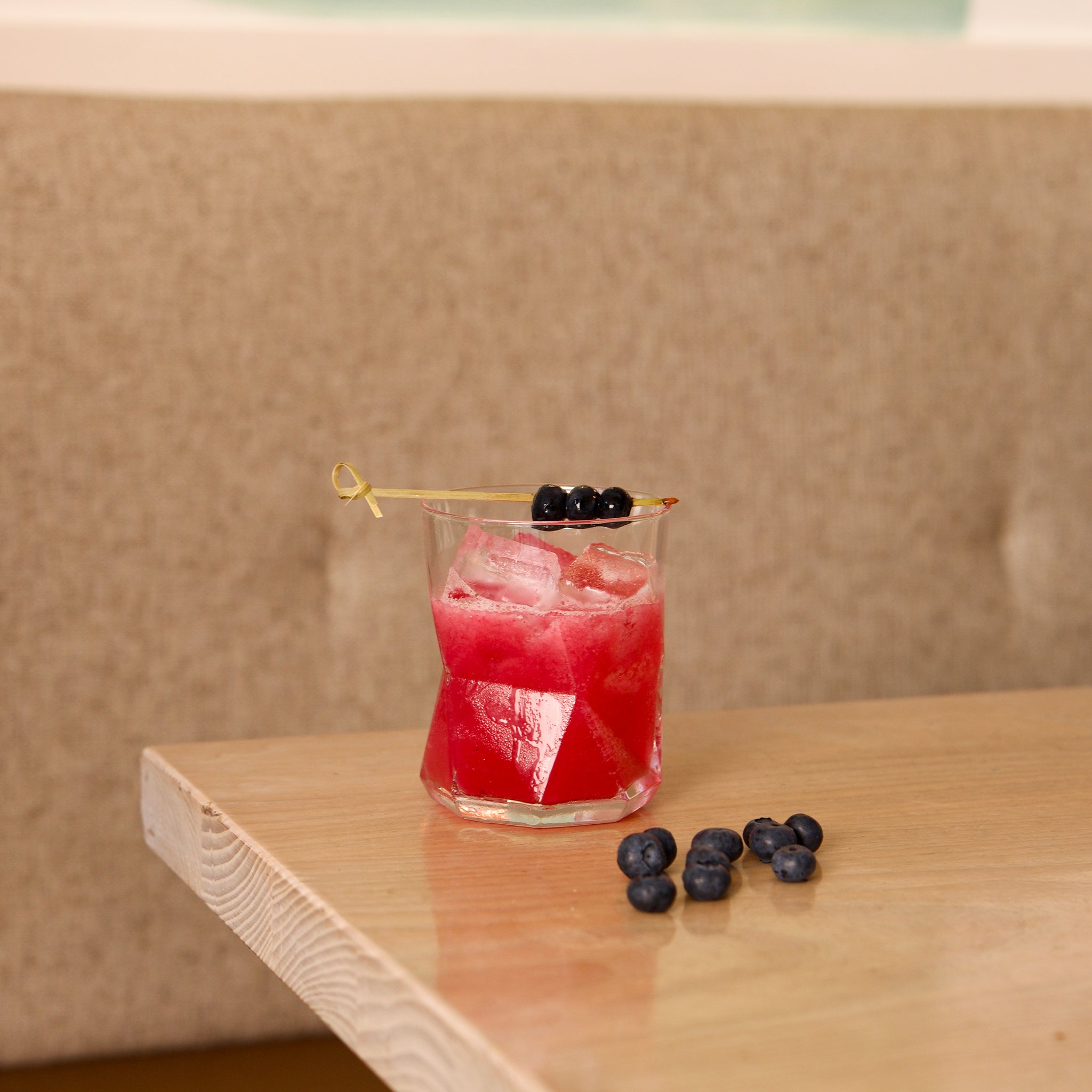 Red cocktail with ice and blueberries in a Cassiopea DOF glass, on a wooden table against a beige wall.