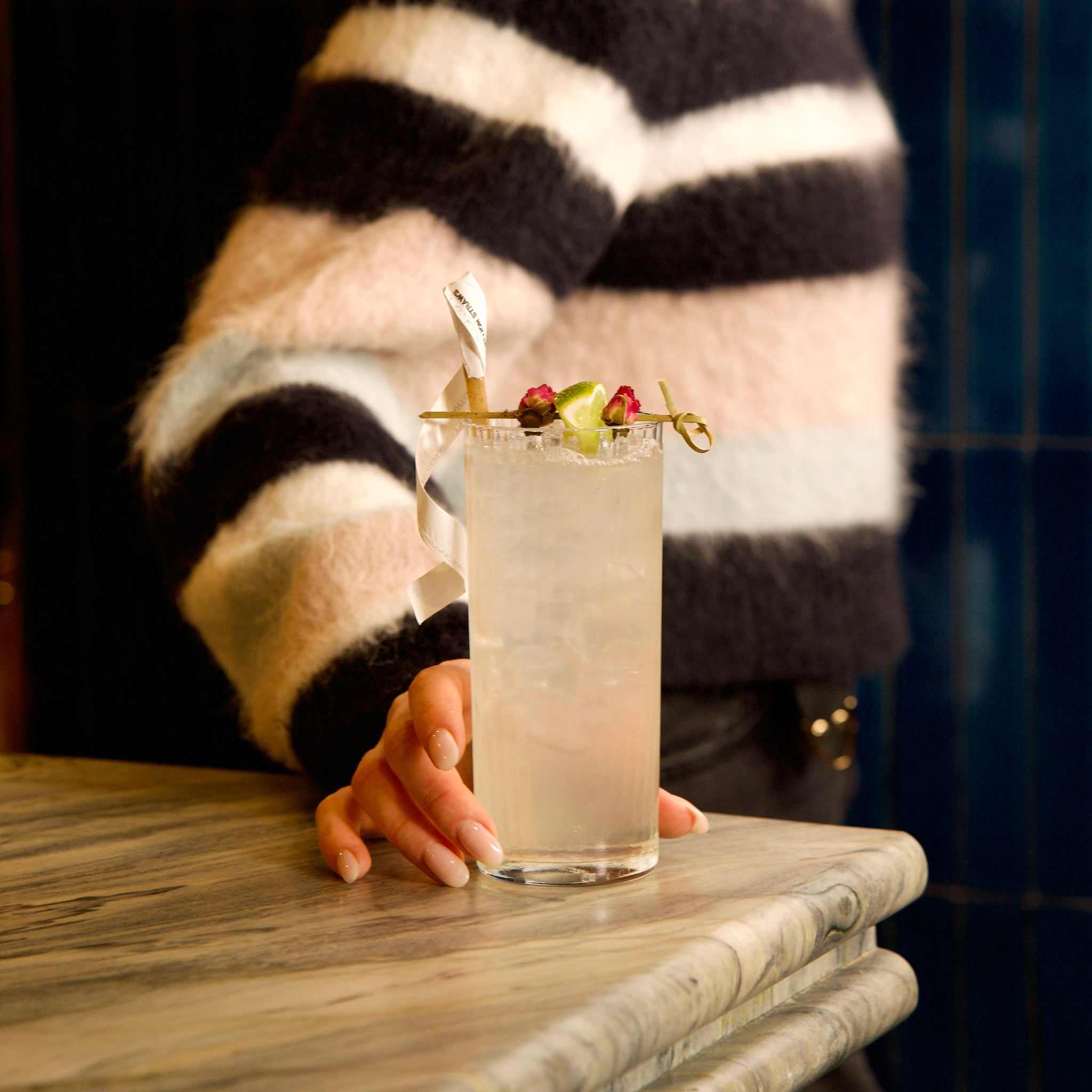 Person in striped sweater holding Exclusiva Tom Collins cocktail glass with a drink with a colorful garnish on a marble surface.