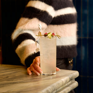 Person in striped sweater holding Exclusiva Tom Collins cocktail glass with a drink with a colorful garnish on a marble surface.