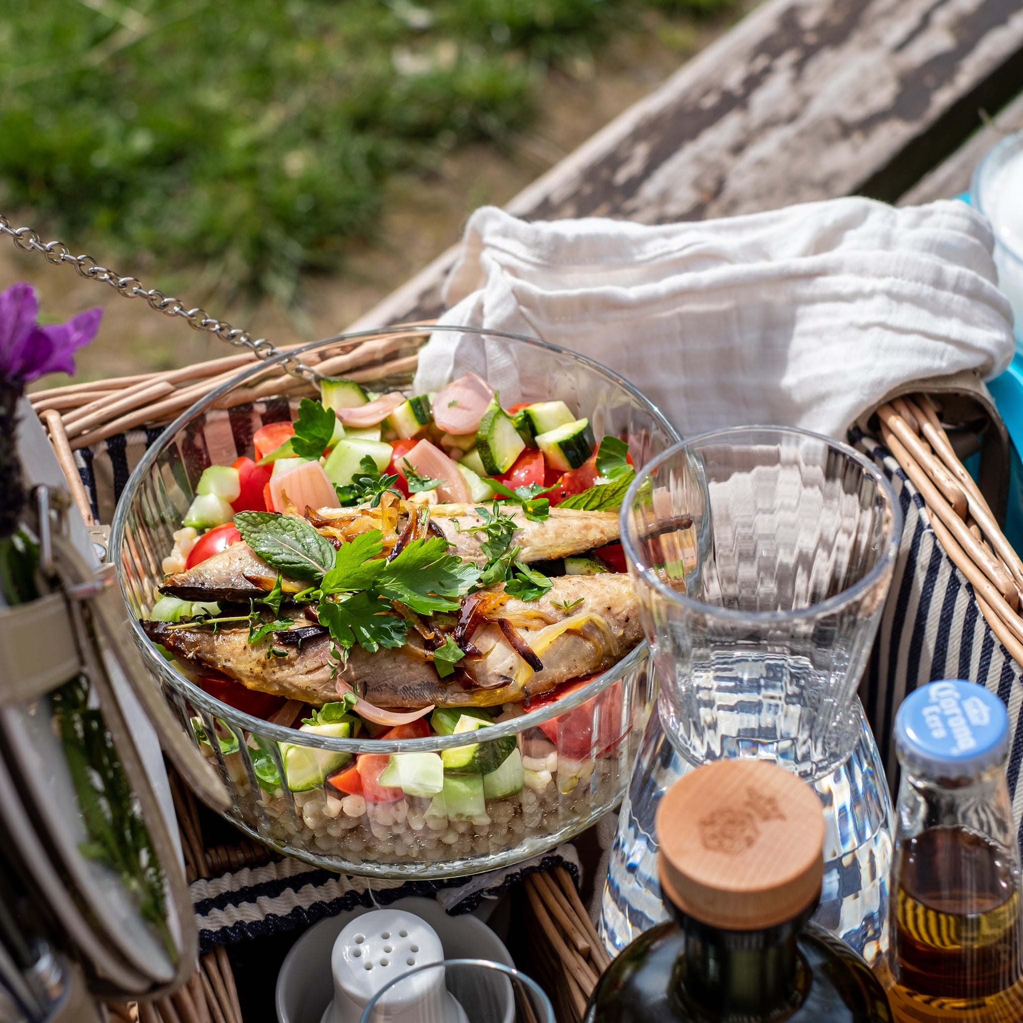 Picnic basket with salad in a Frigoverre Brillante bowl, drinks, and utensils on a grassy background.