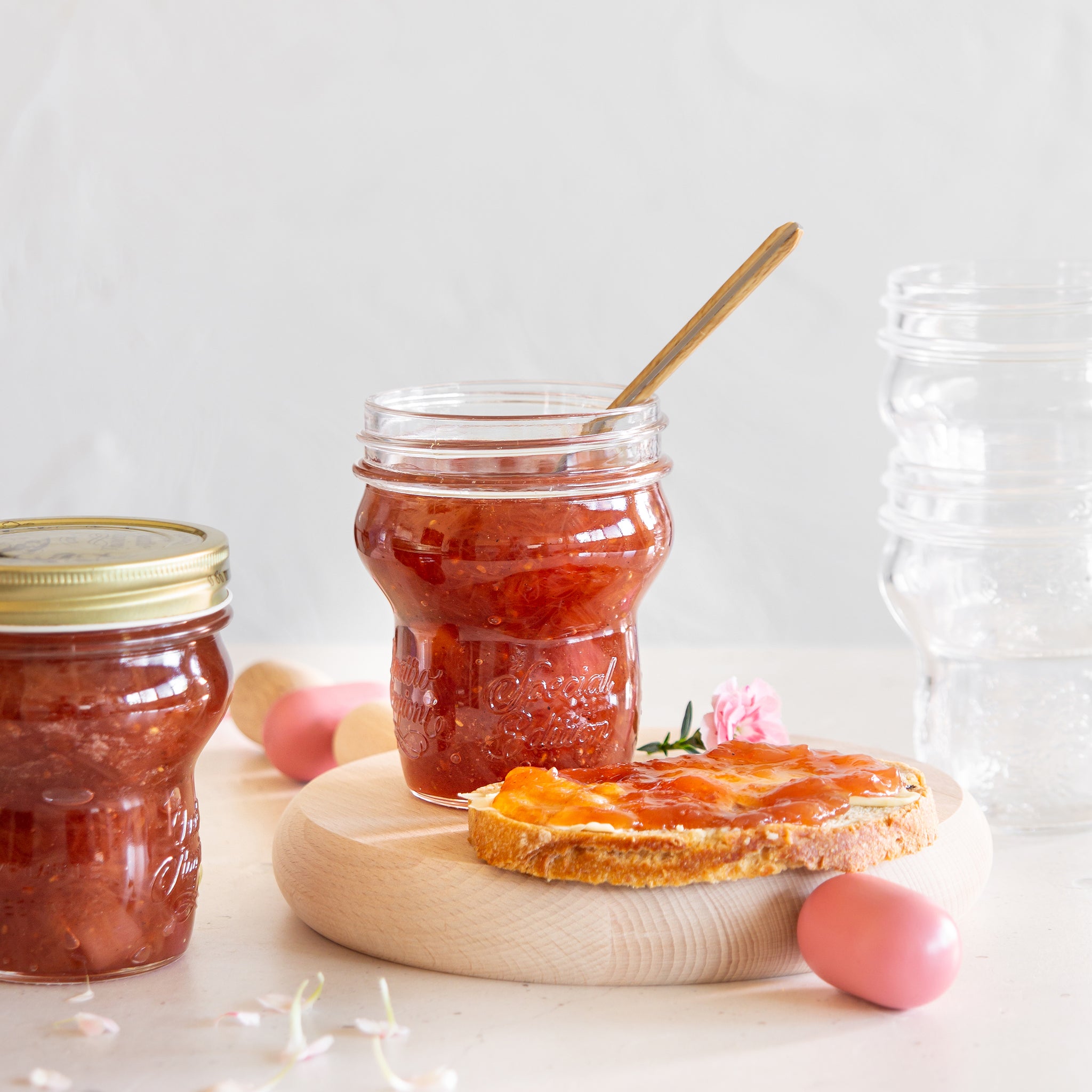 Two Quattro Stagioni special edition jars with red jam with a spoon on a wooden board with a bread slice.