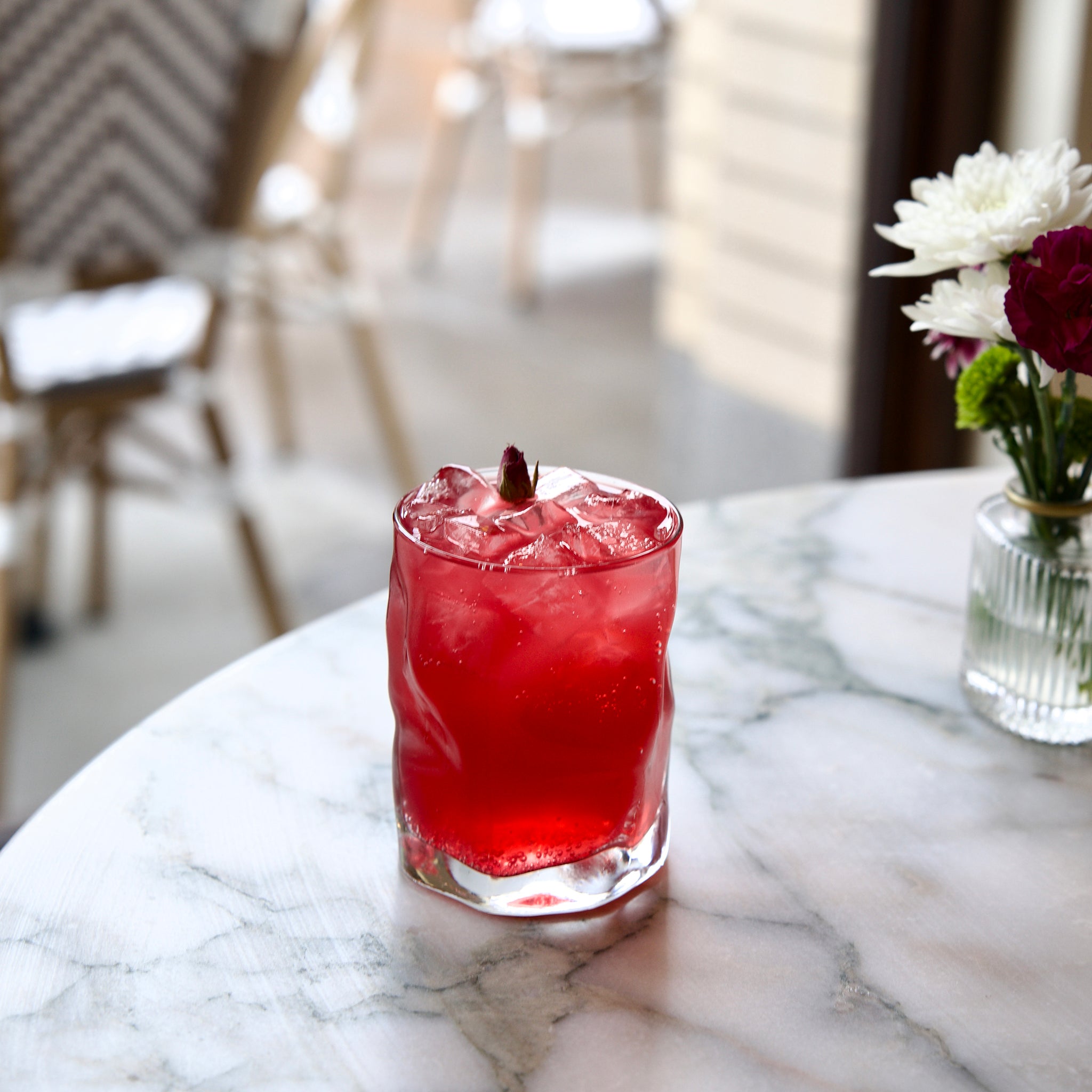 Red cocktail with ice in a Sorgente DOF glass and a garnish on a marble table, with flowers in the background.